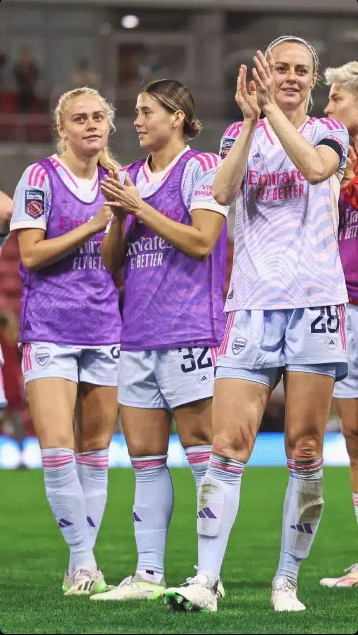 Three Arsenal Women players celebrate after a match, wearing team kits and clapping.