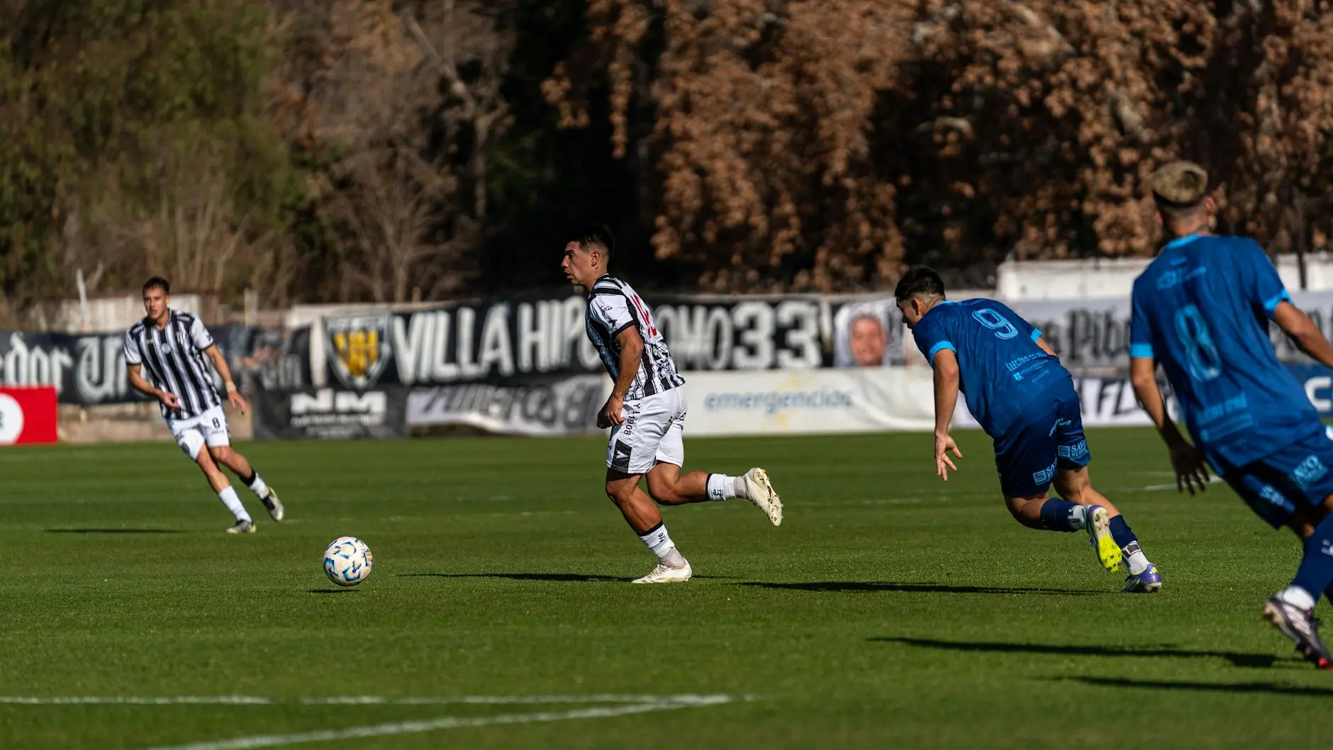 A footballer in a striped jersey sprints with a ball on the pitch, two opponents in blue chase.