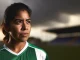 Female soccer player in green kit looking toward horizon with determined expression amid stadium backdrop