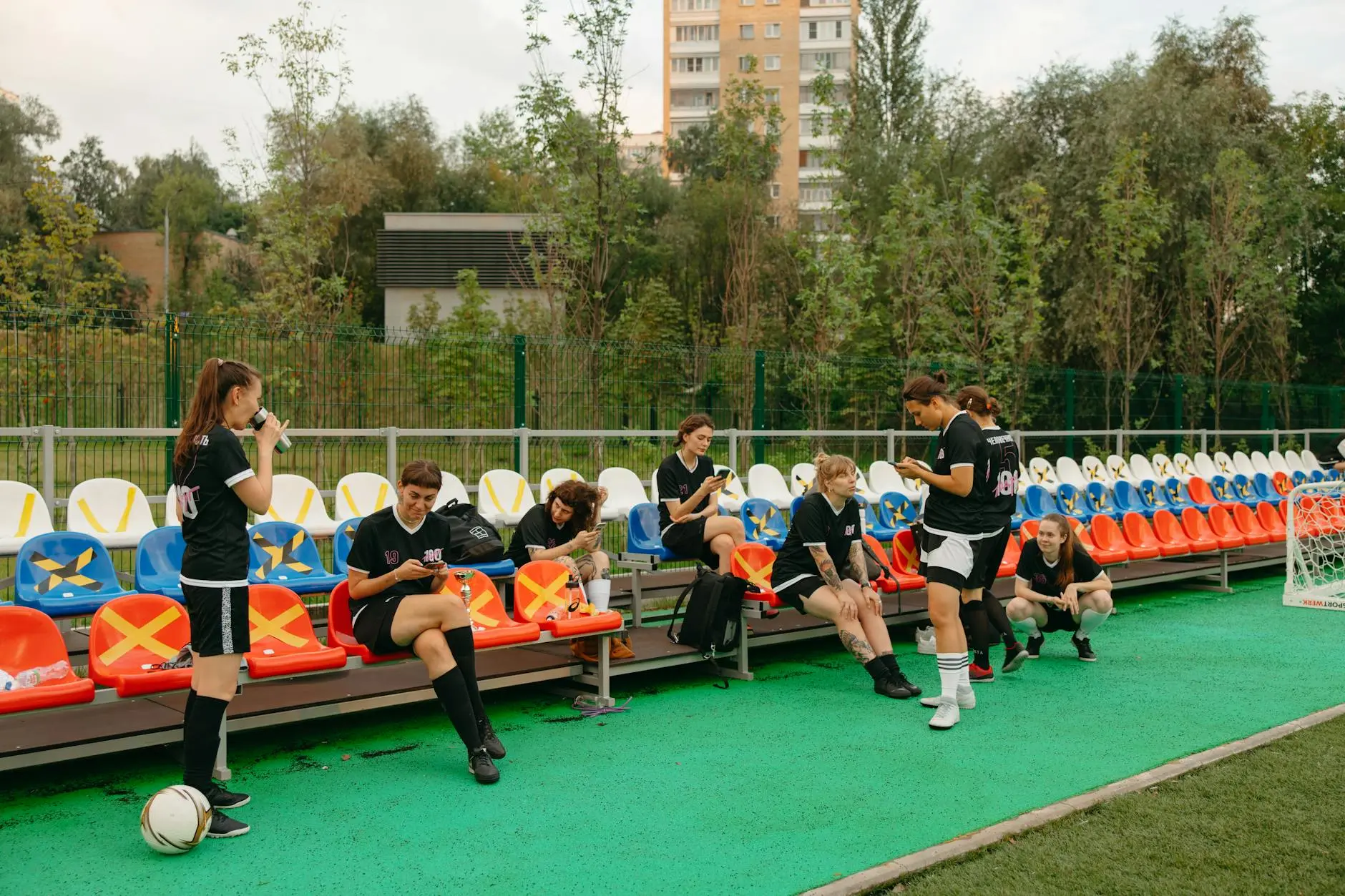 Women football players in training gear sitting on a bench at a pitch, some using mobile devices.