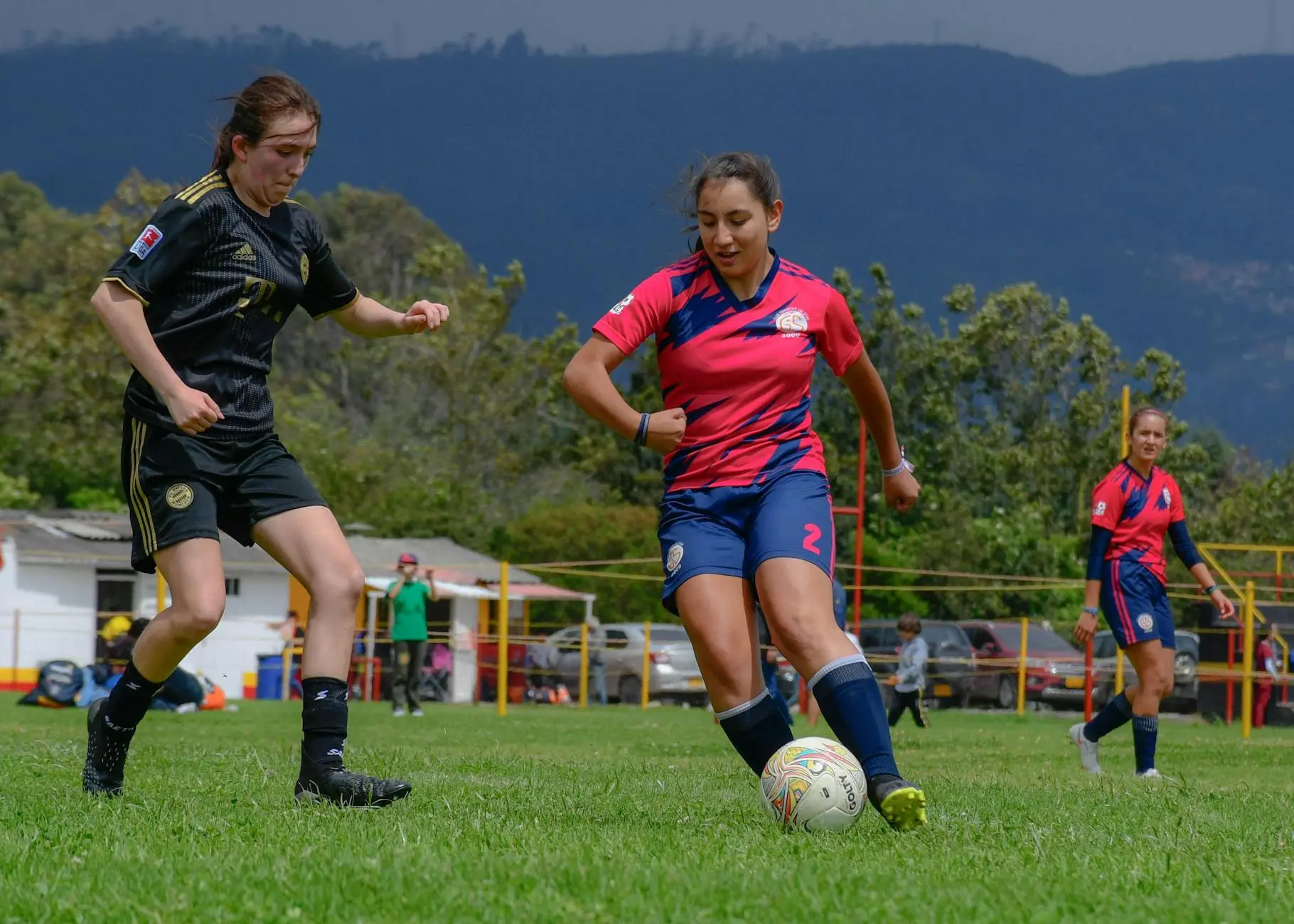 Two female soccer players competing on a grassy field with spectators in the background.