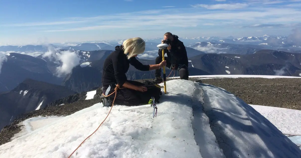 Two people conducting research on a glacier with mountains and clouds in the background.