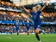 Female footballer in Chelsea kit celebrating goal in stadium with dramatic lighting