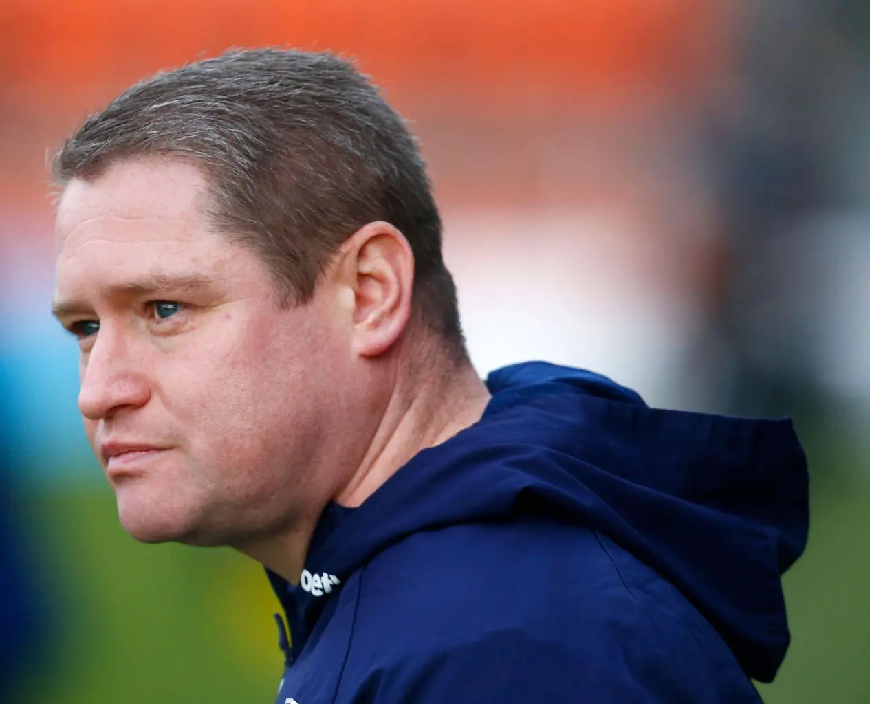 Close-up profile of a man with short hair, wearing a navy jacket, looking thoughtfully.