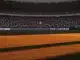Floodlit football stadium at sunset with packed stands, dramatic shadows across empty pitch, crowd lit in green and white.