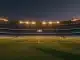 Floodlit stadium pitch at dusk with packed stands, golden-hour lighting, dramatic shadows, no players visible.