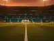 Empty floodlit football pitch at golden hour with long shadows, aerial wide-angle stadium view.