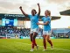 Two Manchester City women footballers celebrating on pitch in sky blue jerseys during golden hour