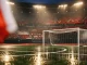 Empty floodlit stadium pitch at dusk with crimson and white light, corner flag, goal net, and tiered stands in soft focus.
