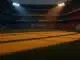 Floodlit football stadium at golden-hour with dramatic shadow patterns across empty pitch, national colours ambient lighting.