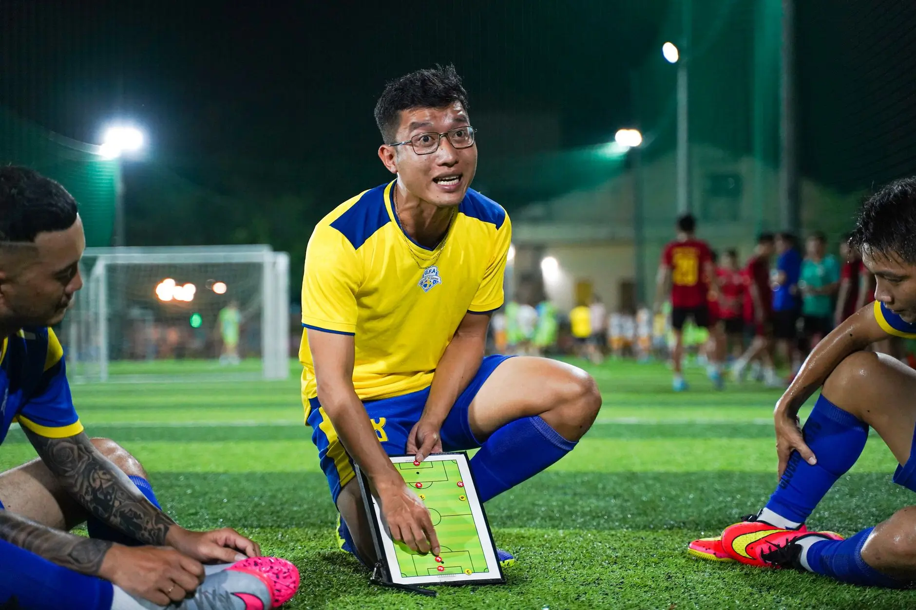 Coach in yellow uniform explaining tactics on a tablet to athletes on a football field.