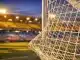 Floodlit football stadium with yellow and blue lighting, empty pitch, billowing goal net in foreground, evening atmosphere.