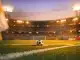 Empty international stadium pitch at dusk with match ball on centre spot, floodlit in orange and yellow-blue stadium lighting.