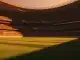 Floodlit stadium at golden hour with packed stands, empty pitch, dramatic shadows, no people or players visible.