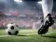 Floodlit stadium pitch with red and white lighting, football boot in motion, ball in centre circle, dramatic shadows.