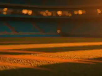 Floodlit stadium pitch at golden hour with turquoise and orange colours, dramatic shadows, packed stands blurred behind.