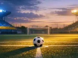 Floodlit football stadium at golden hour with turquoise and yellow lighting over empty pitch and match ball on centre spot.