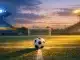 Floodlit football stadium at golden hour with turquoise and yellow lighting over empty pitch and match ball on centre spot.