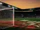 Floodlit football pitch at dusk with red-and-white lit goal net and match ball on centre spot.