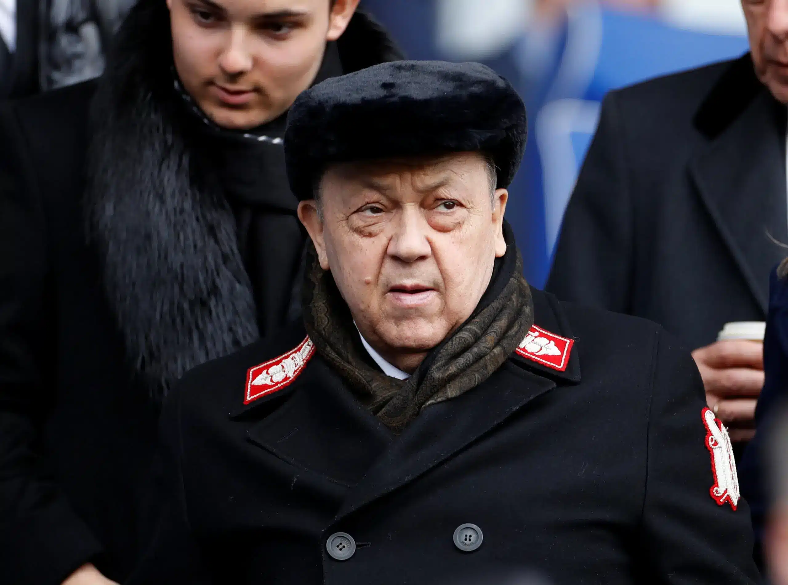 An older man wearing a black fur hat and coat, looking concerned at a football match.