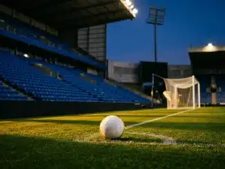 Floodlit stadium pitch at dusk with white football on penalty spot, blue stand sections, golden-hour shadows.