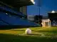 Floodlit stadium pitch at dusk with white football on penalty spot, blue stand sections, golden-hour shadows.