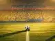 Floodlit stadium pitch with ball on centre spot, bathed in Ecuador-colour lighting—yellow, blue, and crimson tones.