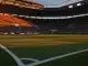 Empty floodlit stadium pitch at dusk with packed stands and dramatic shadow patterns.