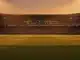 Empty floodlit stadium pitch at golden hour with packed blurred stands in national colours, dramatic shadows and warm evening light.