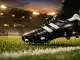 Floodlit football pitch with boot in motion, black and white colour scheme, empty stadium stands at evening light.