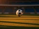 Football mid-flight on floodlit pitch at dusk, dramatic shadows and stadium silhouette, colour-graded in red-gold and blue-white tones.
