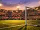 Floodlit stadium stands with colourful bunting and national flag sections at dusk, empty pitch below with dramatic shadows.