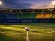 Floodlit stadium with flag-coloured bunting, ball on centre spot, golden evening light casting dramatic shadows on pitch.