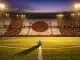 Floodlit stadium at dusk with red and white crowd sections, ball on centre spot, golden evening lighting and dramatic shadows.