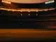 Floodlit stadium at dusk with packed stands and dramatic shadow patterns in amber and blue tones.