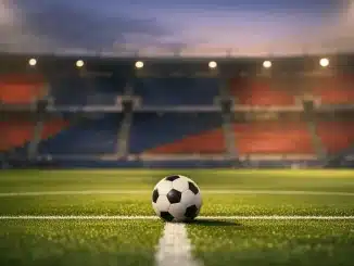 Floodlit football stadium with red and blue stand sections, centre-circle ball under golden spotlight, dramatic shadows on pitch.