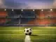 Floodlit football stadium with red and blue stand sections, centre-circle ball under golden spotlight, dramatic shadows on pitch.