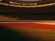 Floodlit stadium pitch at dusk with red-white and green-gold colour grading, dramatic shadows, packed stands in silhouette.