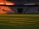 Floodlit football stadium at golden-hour with packed stands, cool green pitch and dramatic amber lighting creating cinematic knockout atmosphere.