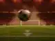 Football pitch under floodlights at evening kick-off, player's boot striking ball in motion, red and green stadium lighting.