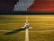 Empty floodlit stadium pitch at dusk with ball on centre spot, red and white seat sections, anticipatory pre-match atmosphere.