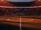 Floodlit stadium pitch at dusk with dramatic shadows, empty turf, packed stands in silhouette, golden-hour mood.
