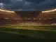 Empty floodlit stadium pitch at dusk with dramatic shadow patterns and golden-hour lighting.