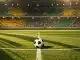 Stadium floodlit in South African flag colours (emerald and gold) with football on centre circle, dramatic shadow patterns, evening kick-off atmosphere.