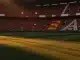 Floodlit stadium at sunset with packed crowds in Spanish and Austrian flag colours, empty pitch ready for match.