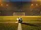 Empty floodlit stadium with football on penalty spot, blue and amber lighting, shadow geometry on pitch.