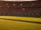 Floodlit football stadium at golden hour with packed stands, emerald pitch in focus, dramatic shadows, Swiss red and Bosnian blue colour palette.