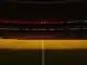 Floodlit stadium pitch at golden-hour kick-off with packed stands in silhouette, warm amber lighting on the field.