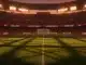 Floodlit stadium pitch at dusk with packed stands, dramatic lighting and shadow patterns, red and navy colour palette.