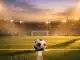 Floodlit football pitch at dusk with ball on centre spot, dramatic shadows and empty stands in soft focus.